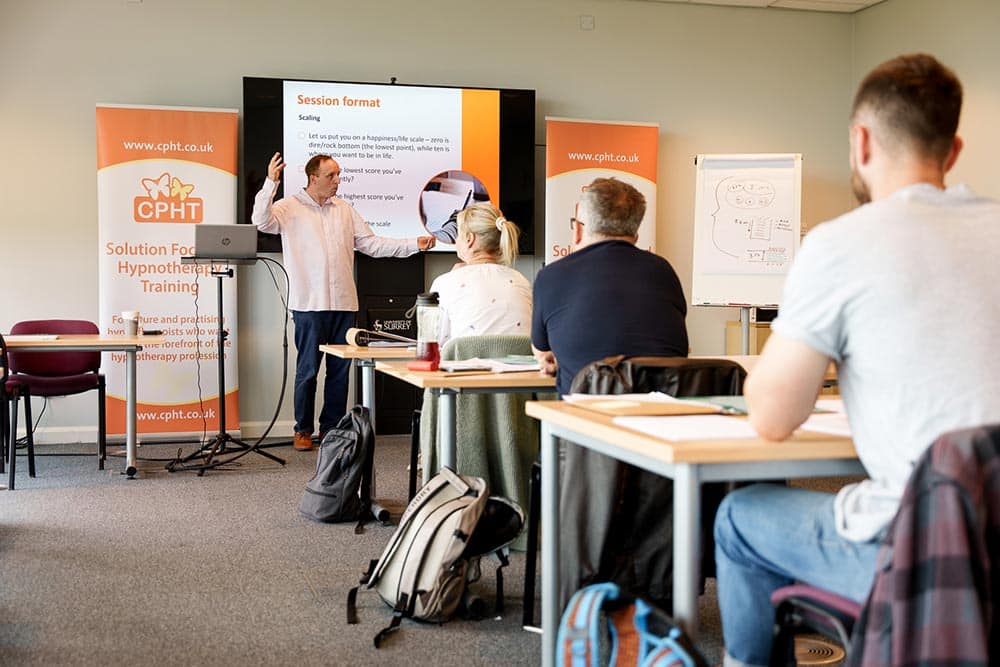 Advanced hypnotherapy certification training classroom with diverse healthcare professionals practicing techniques, instructor demonstrating, and educational materials displayed